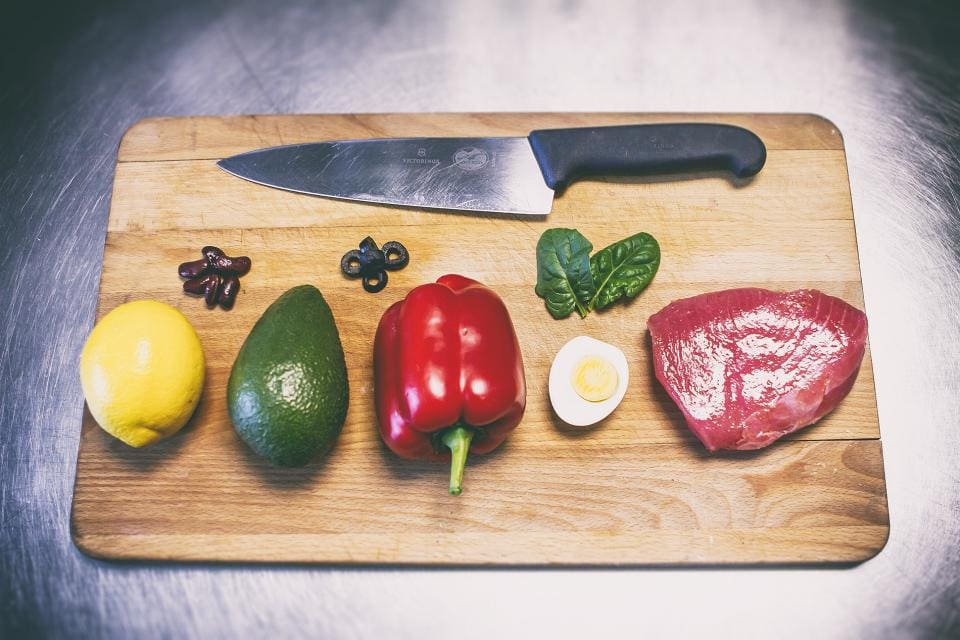 Cooking class: wooden cutting board with large knife on it with cut up olives, and a boiled egg. meat, lemon, avocado and red pepper. 