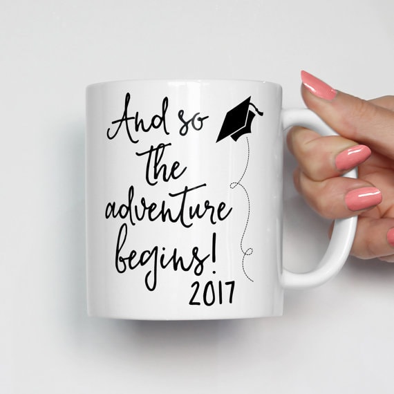 White coffee mug with black font that a flying black cap that says and so the adventure begins and the year of graduation. 