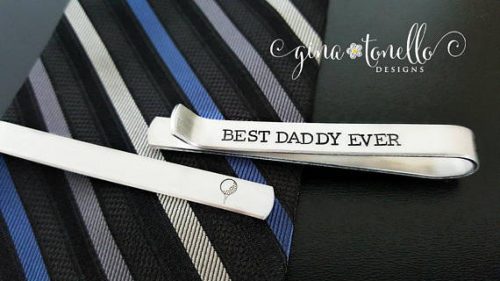 Two silver tie clips on a black, blue, and grey tie, one says Best daddy ever in black text. 