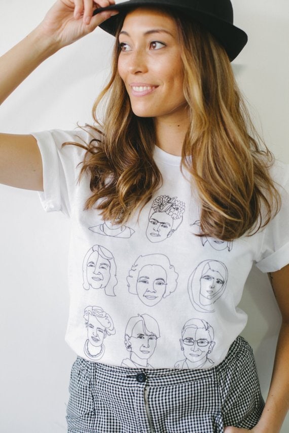 White t-shirt with cartoon style drawings of With Harriet Tubman, Amelia Earhart, Frida Kahlo, Maya Angelou, Malala, and other impactful women on this shirt. 