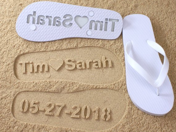Welcome Bag Gifts for Destination Weddings: Sand with a white flip flop right side up and one upside down showing its personalized with Tim hearts Sarah and their wedding date so that it can be imprinted in the sand.