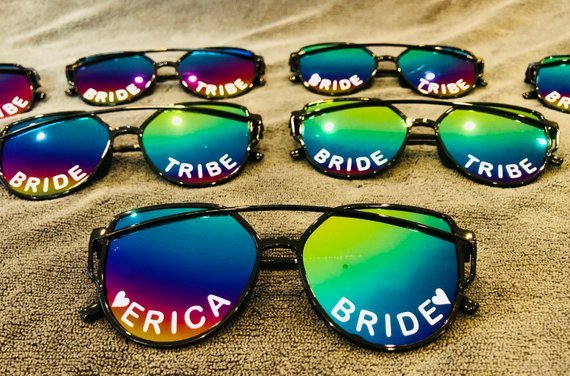 five pairs of customized sunglasses on a sandy beach that says BRIDE TRIBE.