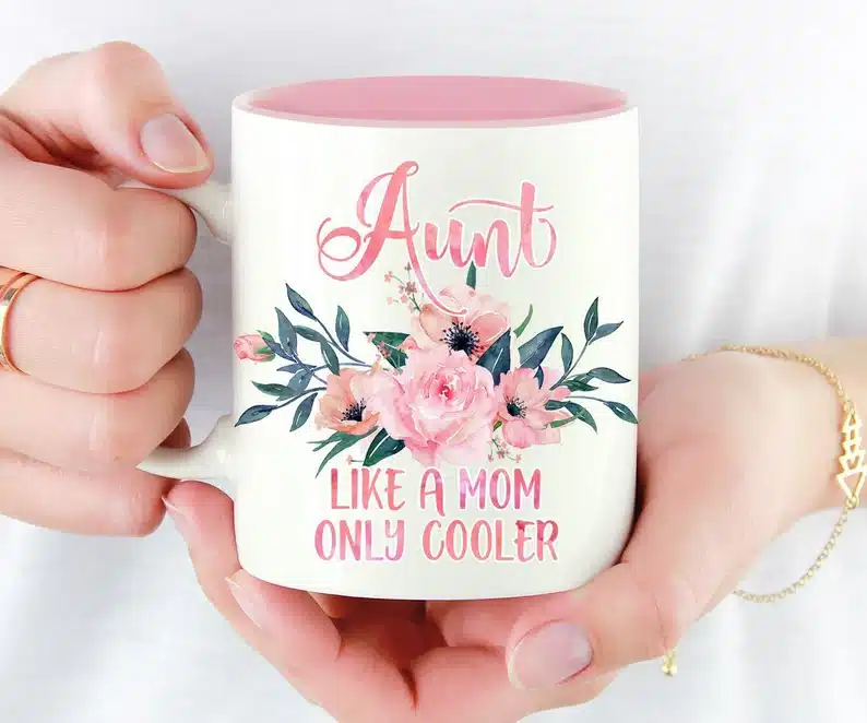 Close up of hands holding a white coffee mug with pink font that says AUNT, with roses below and under the roses more pink font that says like a mom only cooler. 