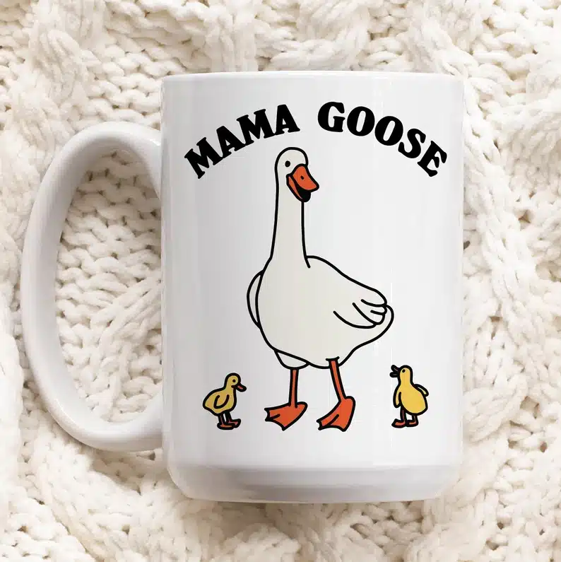 White coffee mug with a tall white goose on it and two little baby yellow chicks with black font above that says MAMA GOOSE. 