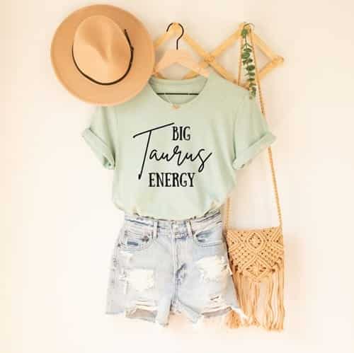 Mint colored t-shirt with black font that says "Big Taurus energy" with a beach hat and beach bag hanging by it.