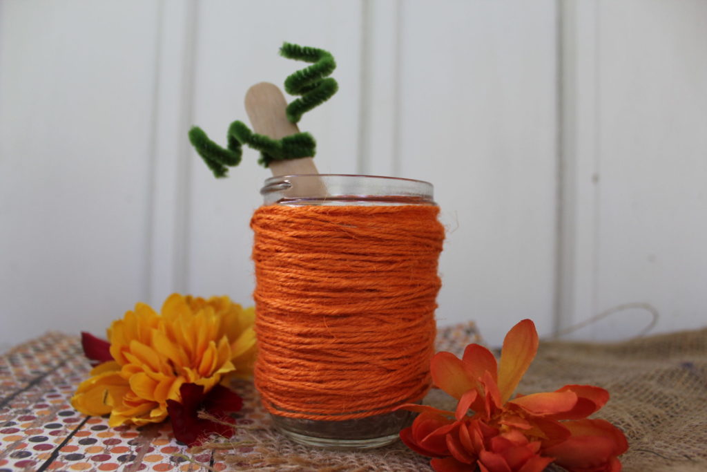 Close up of glass jar covered in orange twine with apopsilce stick and green pipcleaner sticking out of it. 