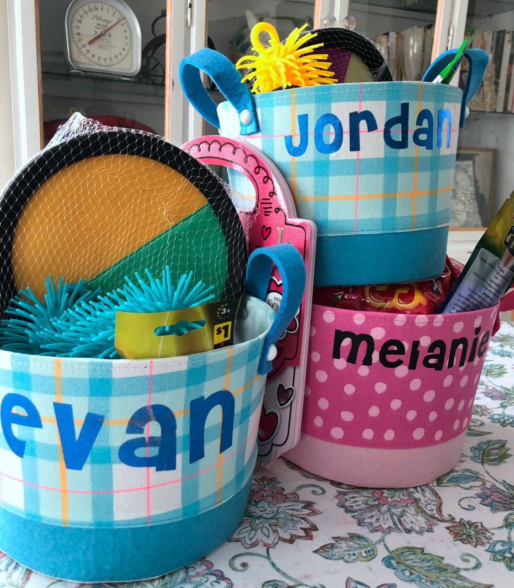 Three baskets with iron on felt names: blue basket with EVAN on it filled with various gifts, pink basket with Melanie on it , and top blue basket with Jordon ironed on. 