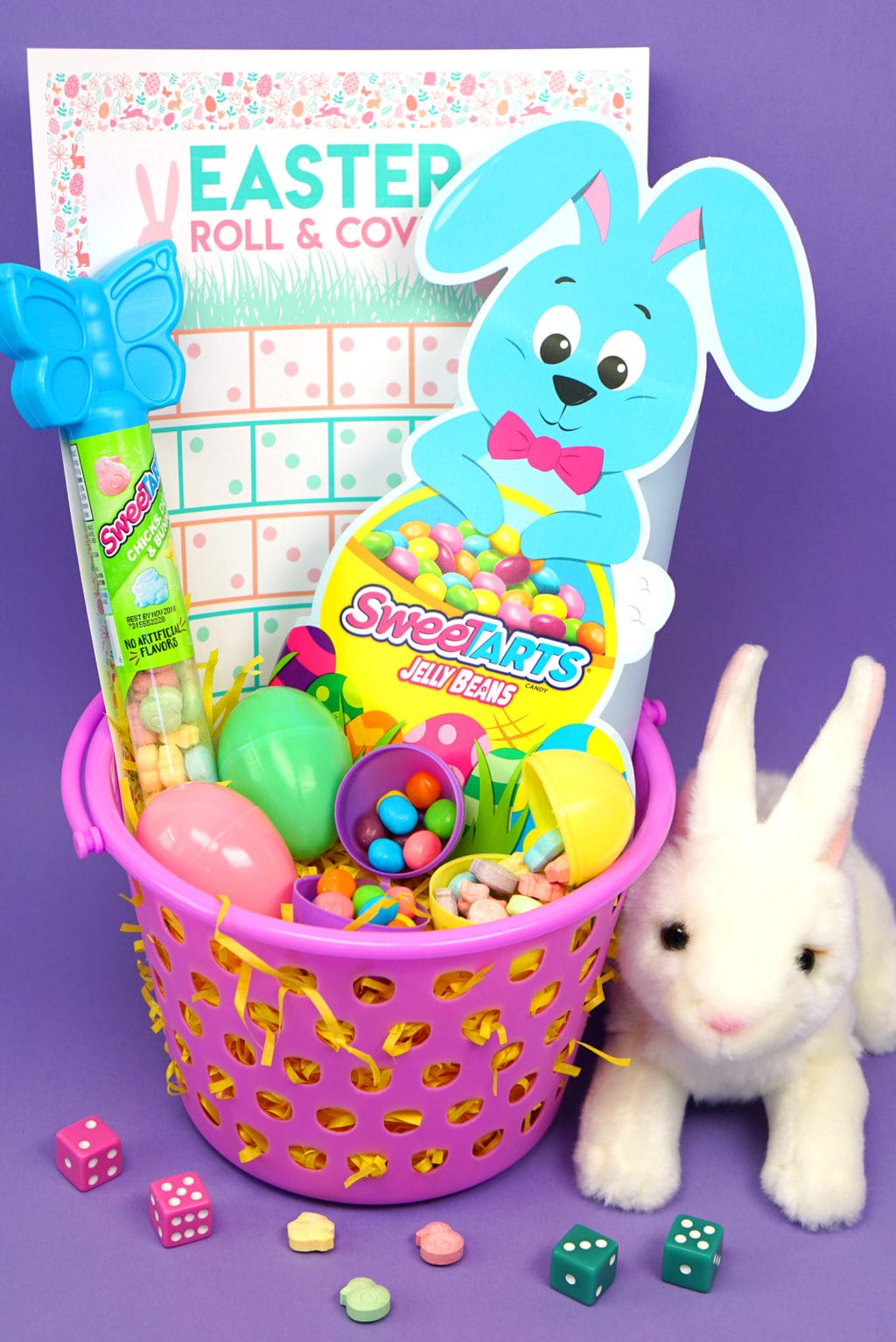Purple Easter basket with various candies in eggs, stuffed white bunny, and Easter themed game. 