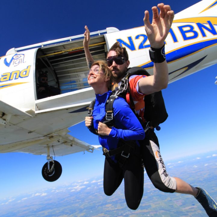 Tandem Skydiving in Indianapolis | A girl in a blue shirt tandem jumping with a guy in a red t shirt, both wearing black pants, with the airplane right behind them, they have just jumped