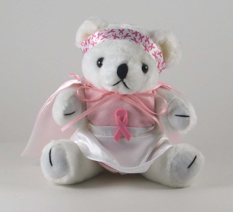 SMall white teddy bear with a pink breast cancer ribbon on its head and a pink outfit.