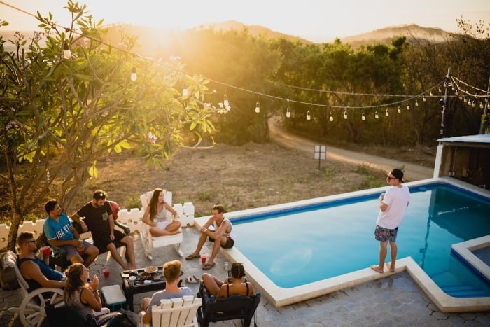 Introvert standing by pool outdoors with group of people in chairs to the right