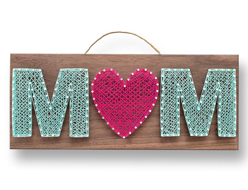 Rectangle wooden sign with string art; teal M, pink heart, and teal M on it. 