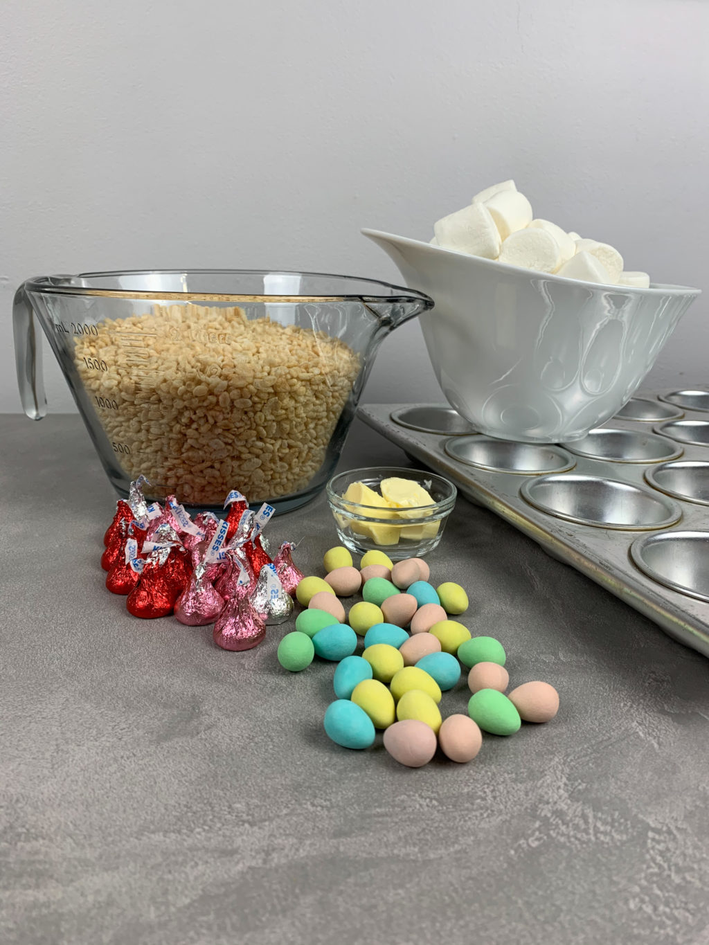 Showing ingredients needed, large glass bowl of rice krispies, various pink and red wrapped heresy kisses, mini eggs loose on the table, small clear glass bowl of butter, muffin tin and a mixing bowl. 