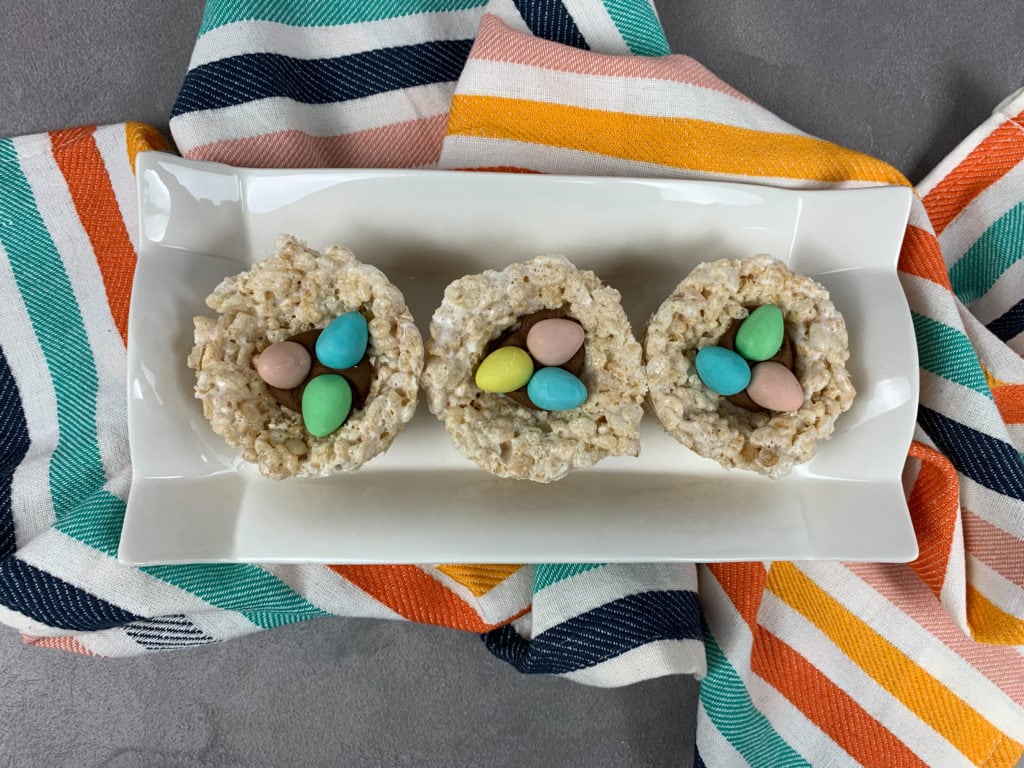 Above view of three completed Rice Krispie Easter Egg Nests on a white rectangle plate on a colorful blanket. 