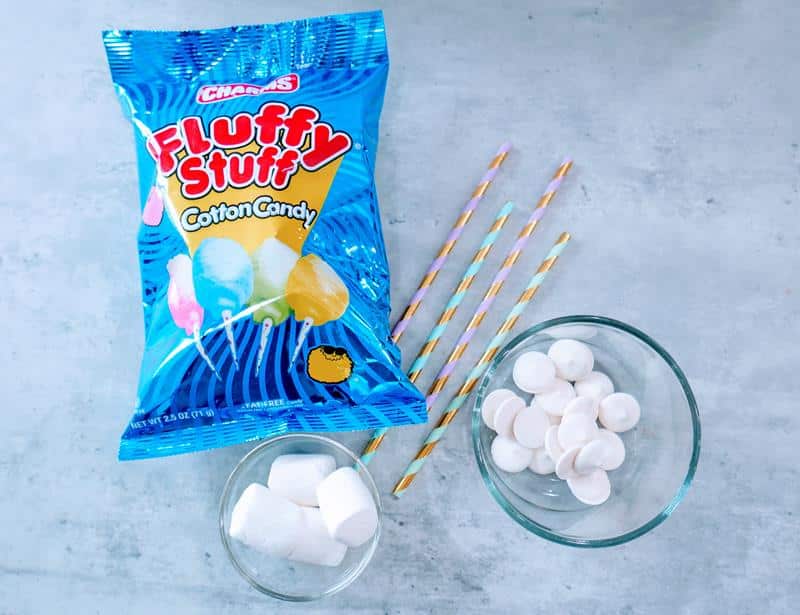 Above view of ingredients required: bag of cotton candy, four gold and silver straws, white candy melts in a glass bowl, and a glass bowl of marshmallows. 