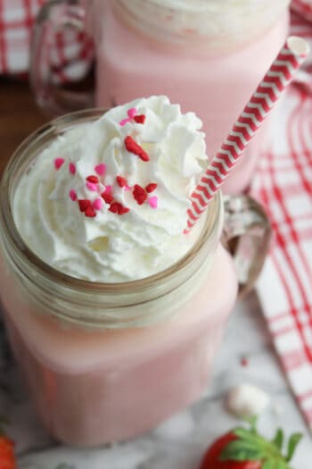 above veiw close up of pink hot chooclate with whipped cream, sprinkles and a cute red and white straw sticking out of a masion jar glass with a handle.