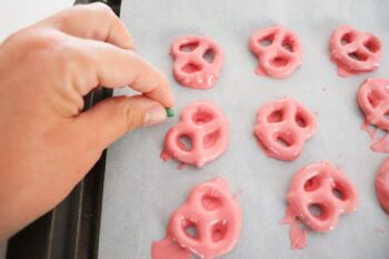 close up of pretzels, showing hand adding green M&M to top of candy melts. 