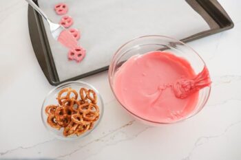 baking sheet with parchment paper on it with 4 completed pretzels covered in candy melts, glass bowl of pretzels and glass bowl of candy melts by baking sheet. 