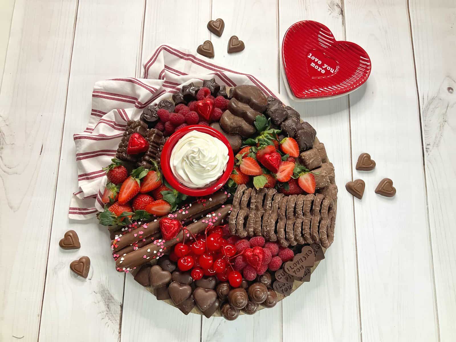 completed chocolate lovers valentines day snack board with cherries, strawberries, raspberries, homemade peanut butter chocolate hearts, chocolate hearts, chocolate covered pretzels, and a bowl of cool whip. 