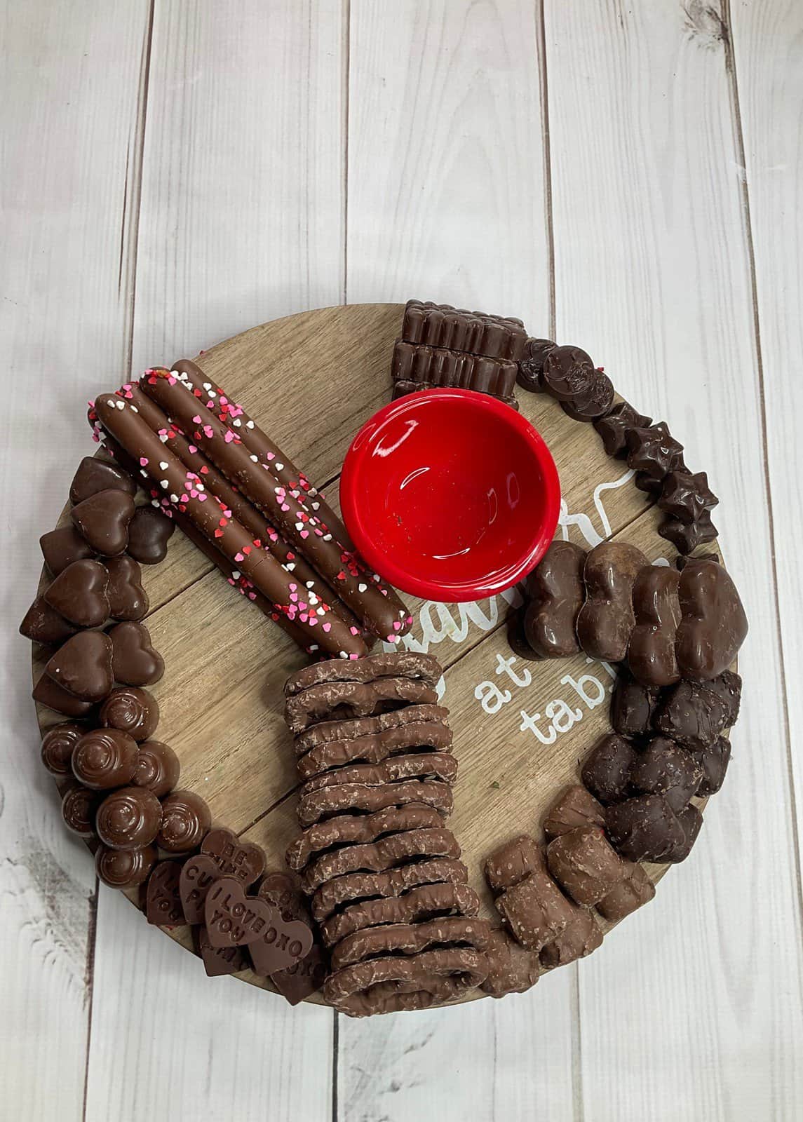 Above view showing round board with chocolate pretzels and hearts on board with red empty bowl.