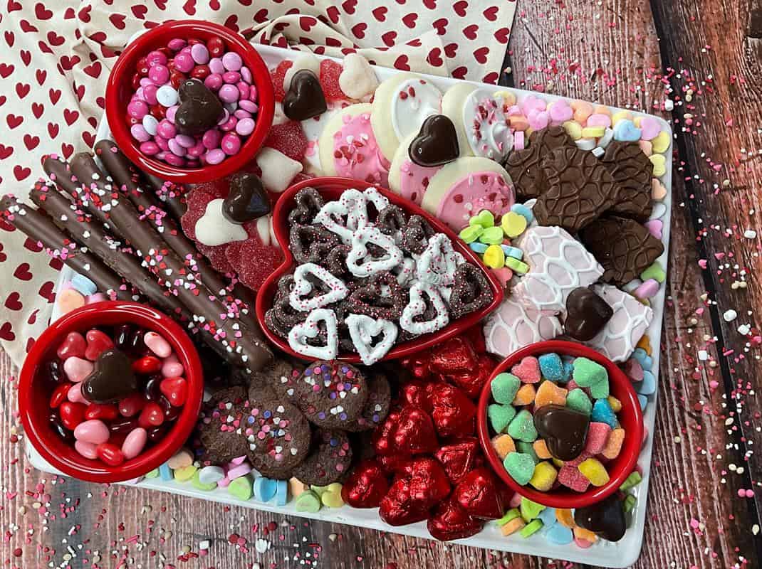 Completed snack board showing little red bowls filled with jellybeans, sugar candy hearts and M&M with pretzels, sugar cookies, brownie's, and various valentine themed treats surrounding them.