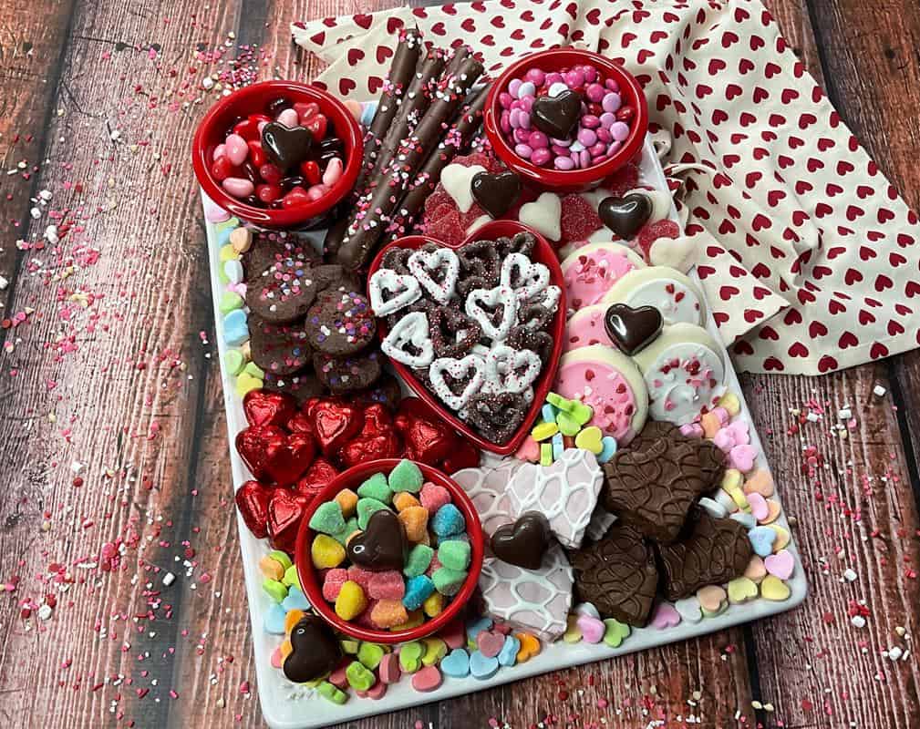Above view on snack board with valentine themed pretzels, M&M, jelly hears, sugar cookies, brownies, jelly candy, little Debbie snack cakes, chocolate hearts, and conversation hearts. 
