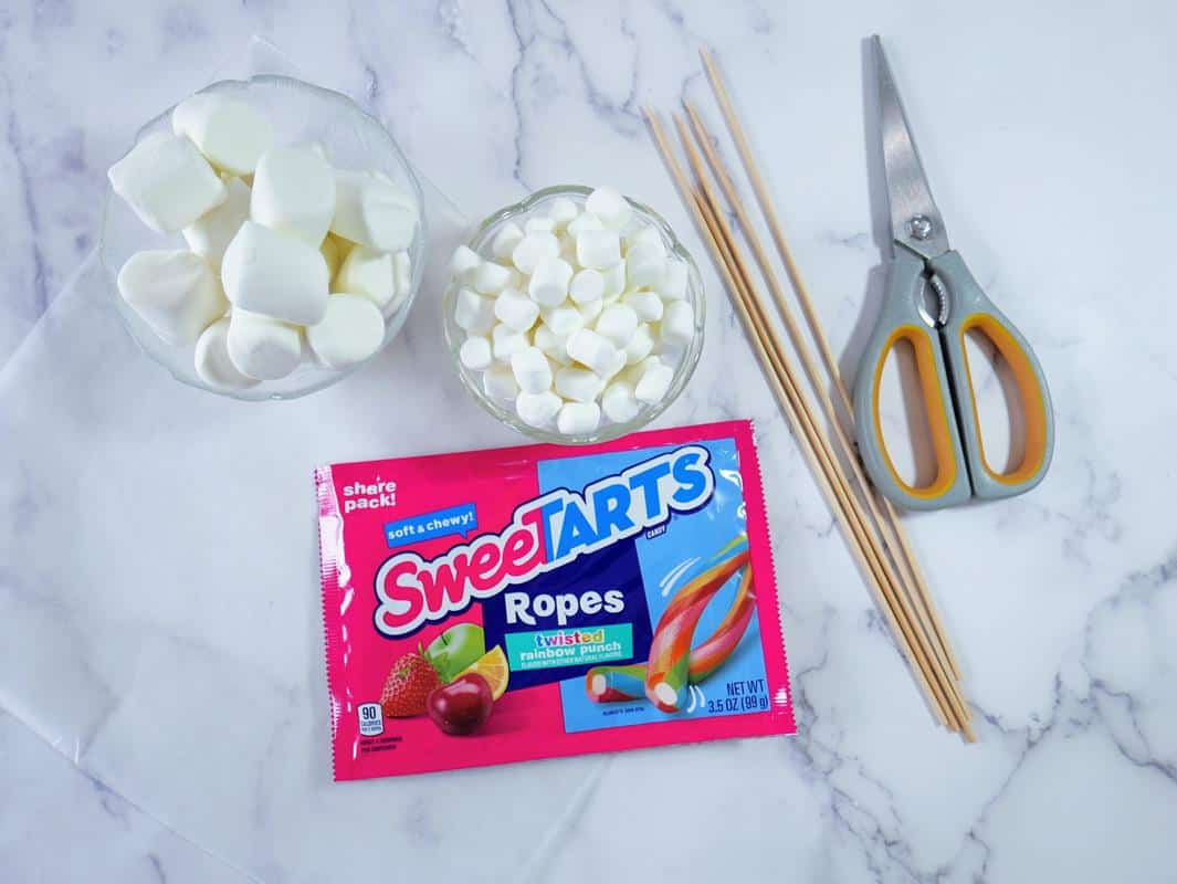 Above view of ingredients required: bowl of large white marshmallows, bowl of mini marshmallows, bag of sweetarts ropes, five bamboo skewers, and scissors. 
