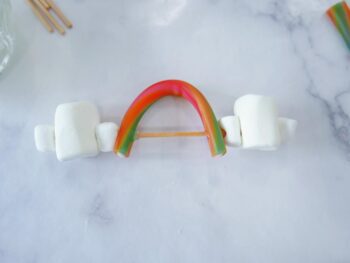 Showing mini marshmallow, regular size marshmallow, mini marshmallow and sweetart candy arched like a rainbow, followed by another mini marshmallow, regular size marshmallow and a mini marshmallow on bamboo skewer