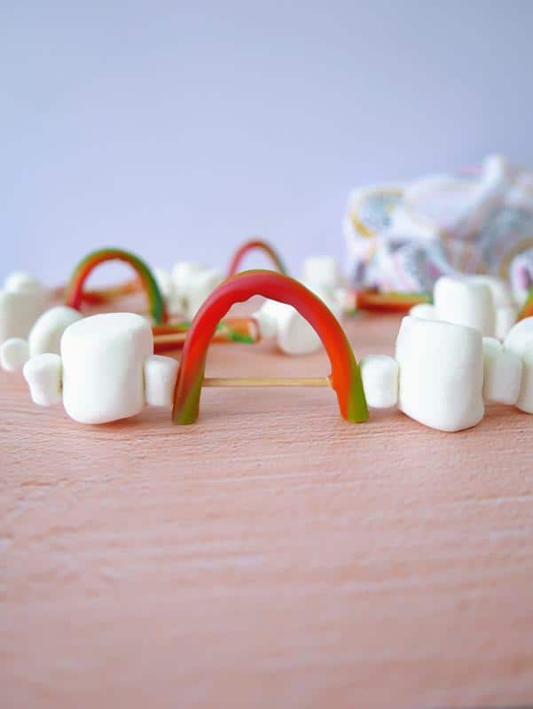 Front view of rainbow skewer snacks, showing bamboo skewer with 3 mini marshmallows one each side of an arched rainbow candy. 