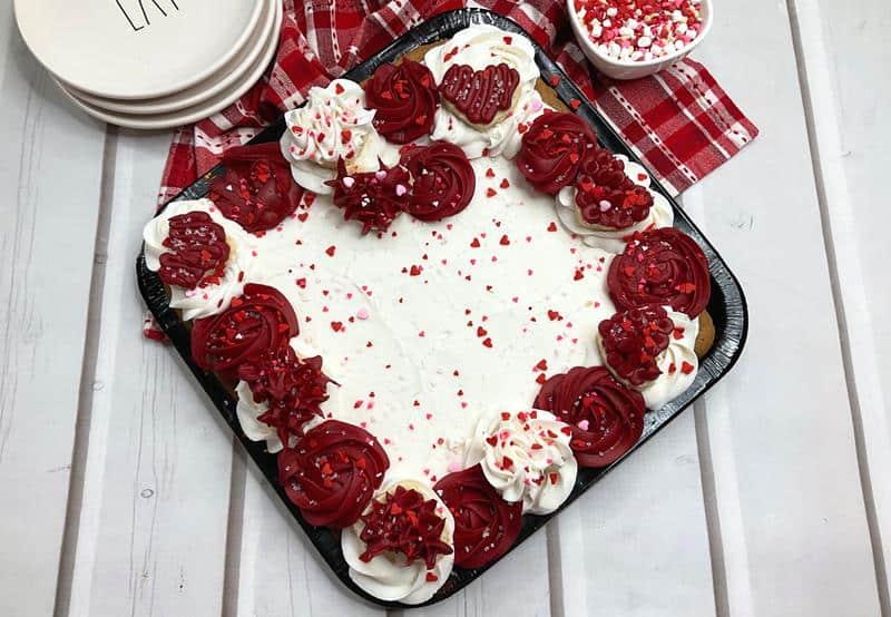 square valentines day cookie cake with red and white frosted roses, heart shaped sugar cookies, and heart sprinkles all over it. 