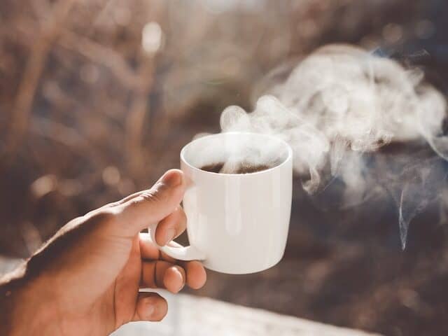 Hand holding a white coffee cup filled with steaming hot coffee. 