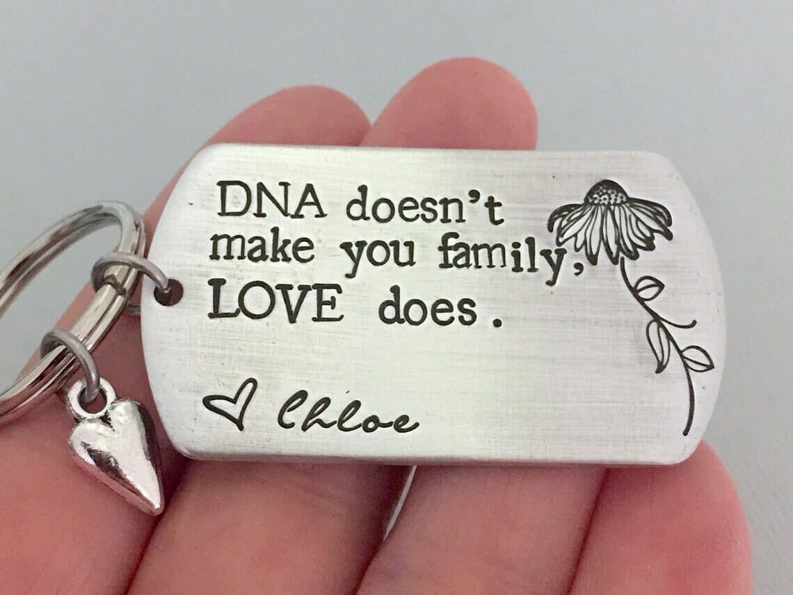 Silver dog tag charm engraved in black that says DNA doesn't make you family, LOVE does. with a flower beside it. 