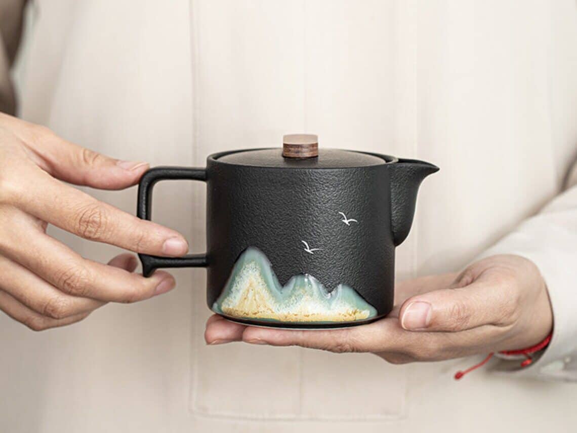 Gift Ideas for an Aries Woman: Person holding a small black teapot with mountains and birds painted on it.