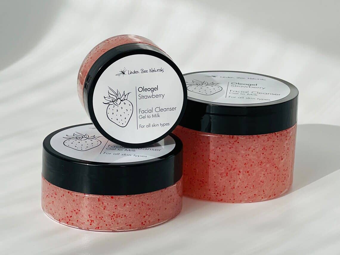 Three small containers clear bottoms that show a pink and darker pink spotted face scrub in them, with black lids and a white top label that says its strawberry. 