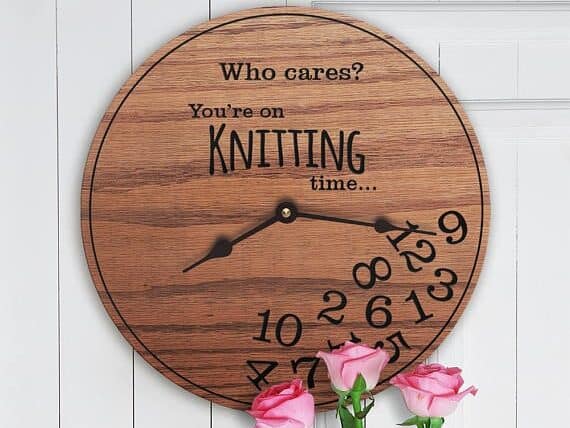 Round wooden clock with black hands, font on it that says Who cares? You're on knitting time... and all the numbers on the clock at the bottom in a pile.