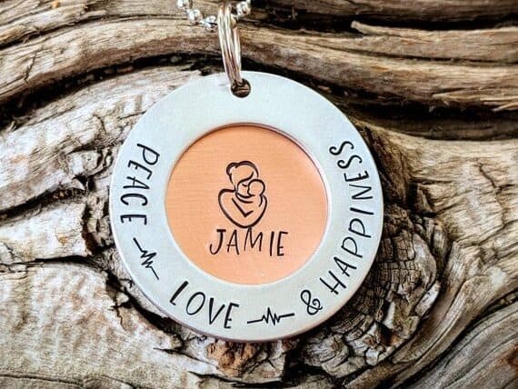 Mother's Day Gift for Your Ex Wife: Round white charm with a mother huffing a baby in the middle drawn on it. With writing that says Peace Love Happiness. 