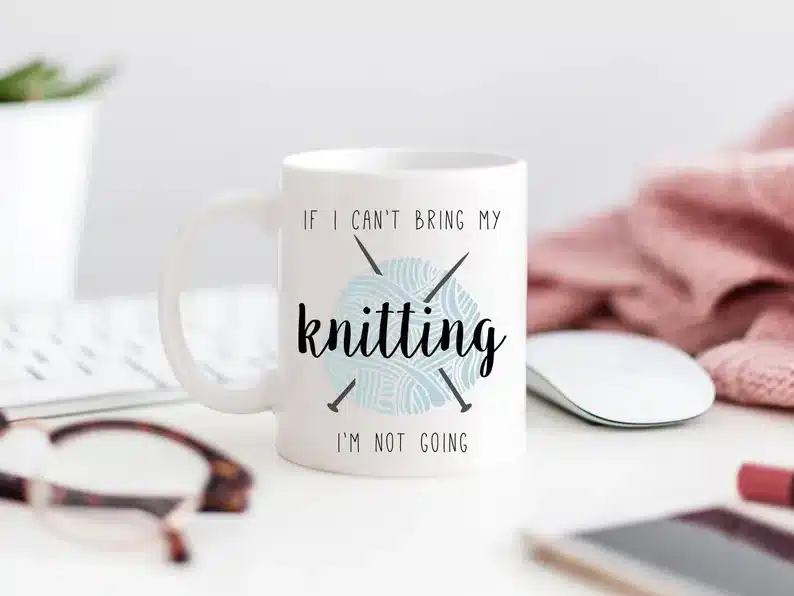 White coffee mug with black font that says If I can't bring my knitting I'm not going. With a blue yarn ball and black needles in the background. 
