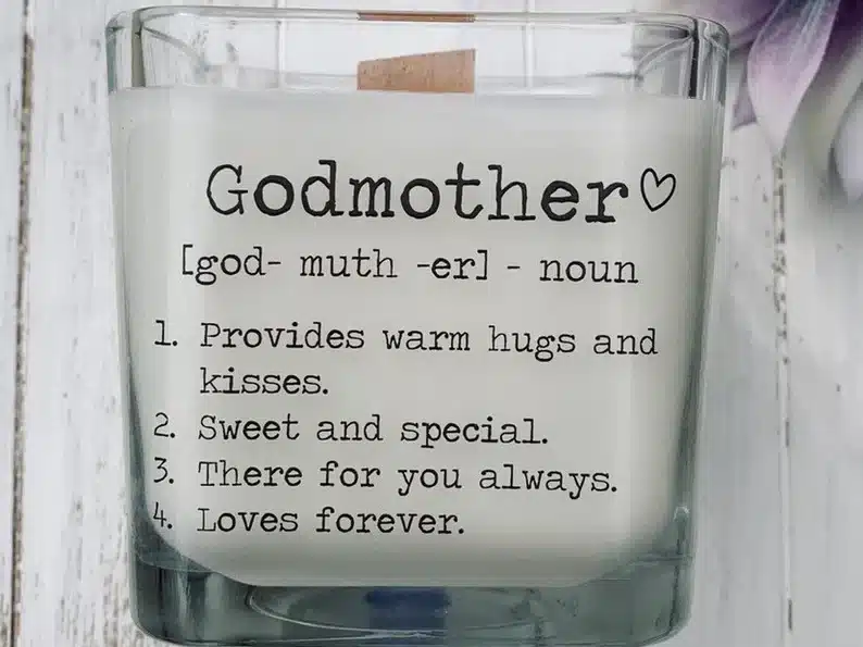 White candle in a clear glass container with black font that says Godmother. 