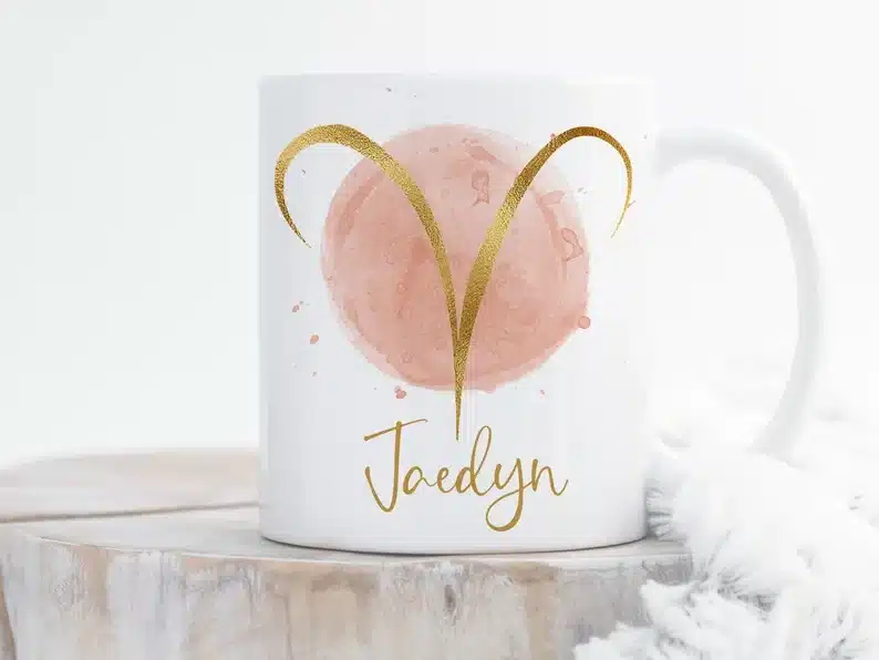 White coffee mug with a pink circle with golden Aries symbol and the name Jaedyn below in gold. 
