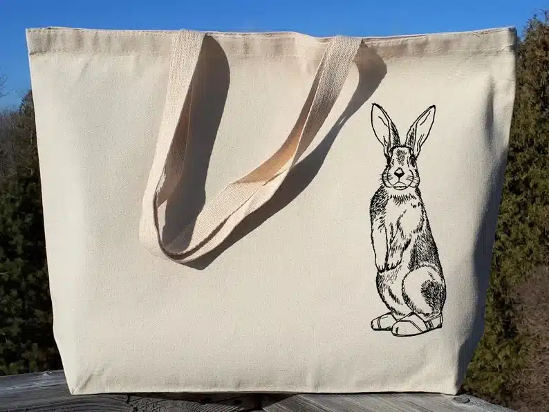 Tan colored tote bag with a black outline of a bunny standing on the right side. 
