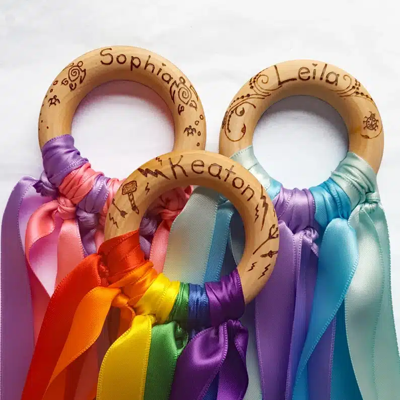 Three rainbow hand satins, all with differnt kids names o the wooden ring with colorful ribbon hanging from them. 