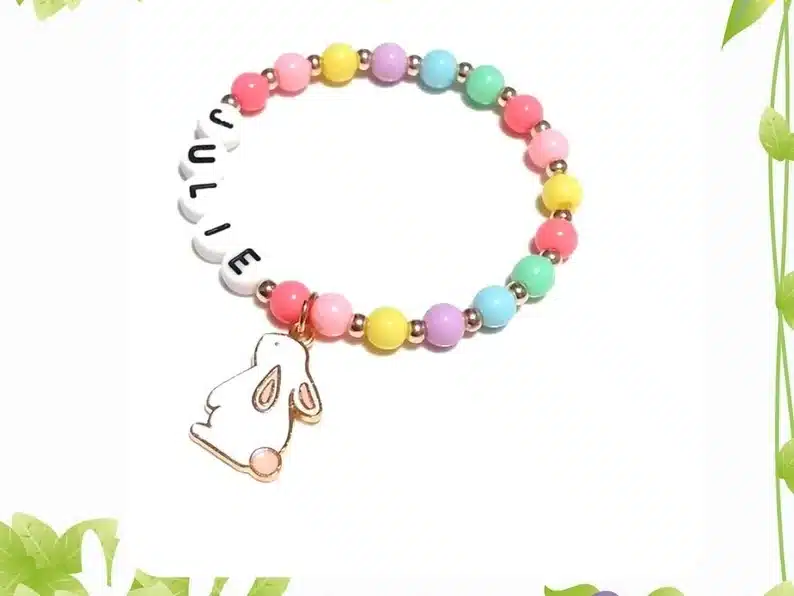 Colorful beaded bracelet: rose pink, pink, yellow, purple, blue, mint green, and repeat around. with gold balls smaller between each bead, with beaded letters that spell JULIE on one side and a white bunny charm hanging from it. 