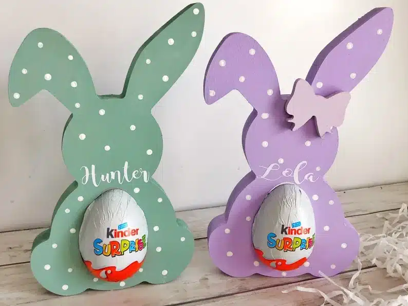 Two customized wooden bunnies, one teal with white dots with the name Hunter in white font and a kinder egg in the middle, the other bunny lavender in color with white dots and white font that says Lola with a kinder egg in it. 