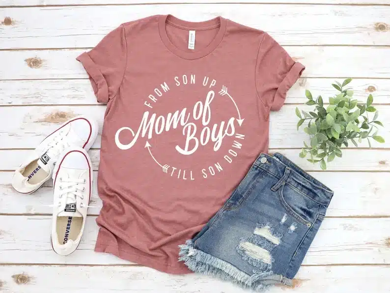 Pink t-shirt laying on white wooden background that has white font that reads "from son up till son down in a circle with MOM OF BOYS in the middle. 