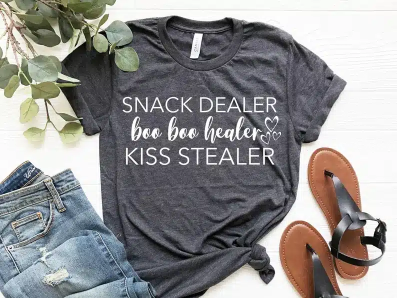 Mother’s Day Gift Ideas for My Wife: Dark grey t-shirt with white font that says snack dealer, boo boo healer, kiss stealer. 