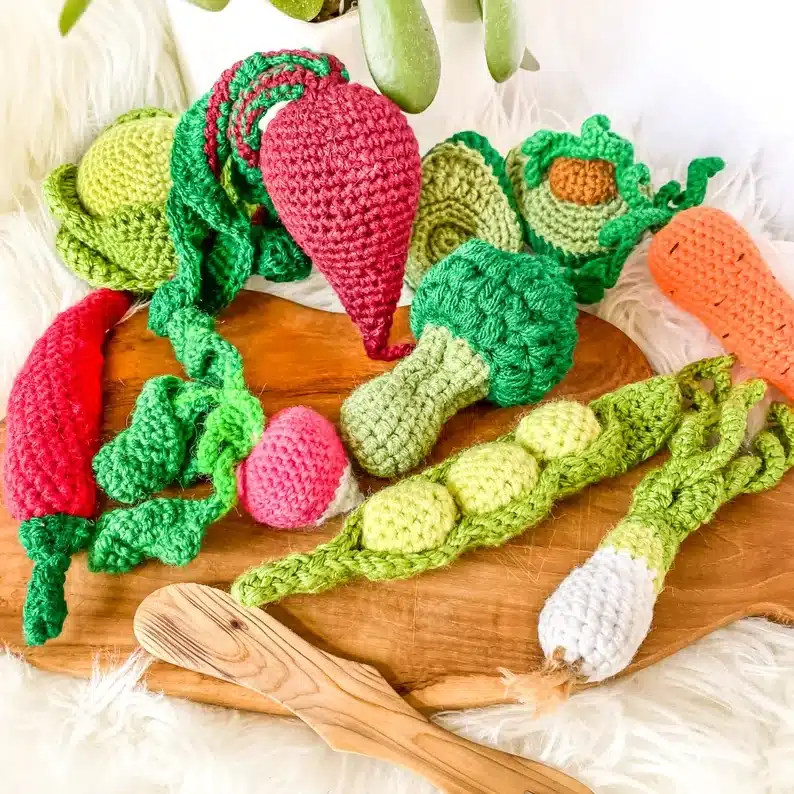 Wooden board with various crocheted garden vegetables; pea pods, parsnip, radish, hot pepper, cabbage, carrot, beet, and avocado. 
