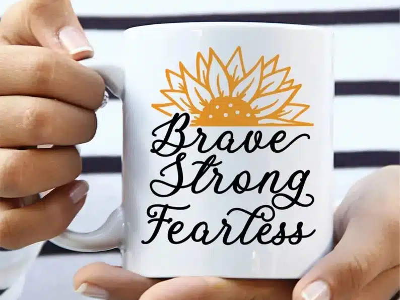 Hand holding a white coffee mug with a yellow half sunflower on the top and black font that says Brave, Strong, fearless below. 