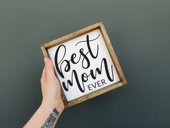 Woman's hand holding a small wooden framed white sign with black font that says Best mom ever. 