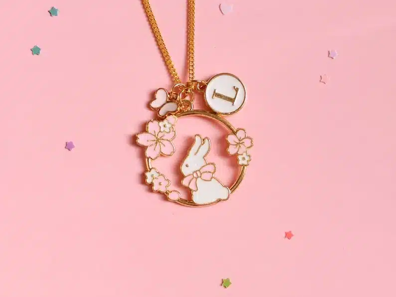 Pink background with a gold nacklace with a circle charm with a white bunny in it with pink flowers around the circle. 