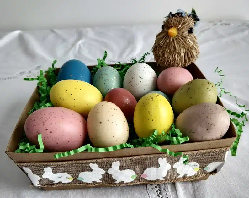 Box with ribbon that has white bunnies all over it, inside box many wooden eggs all differnt colors: pink, tan, yellow, green, blue and white. with a fake bird on the side of the basket. 